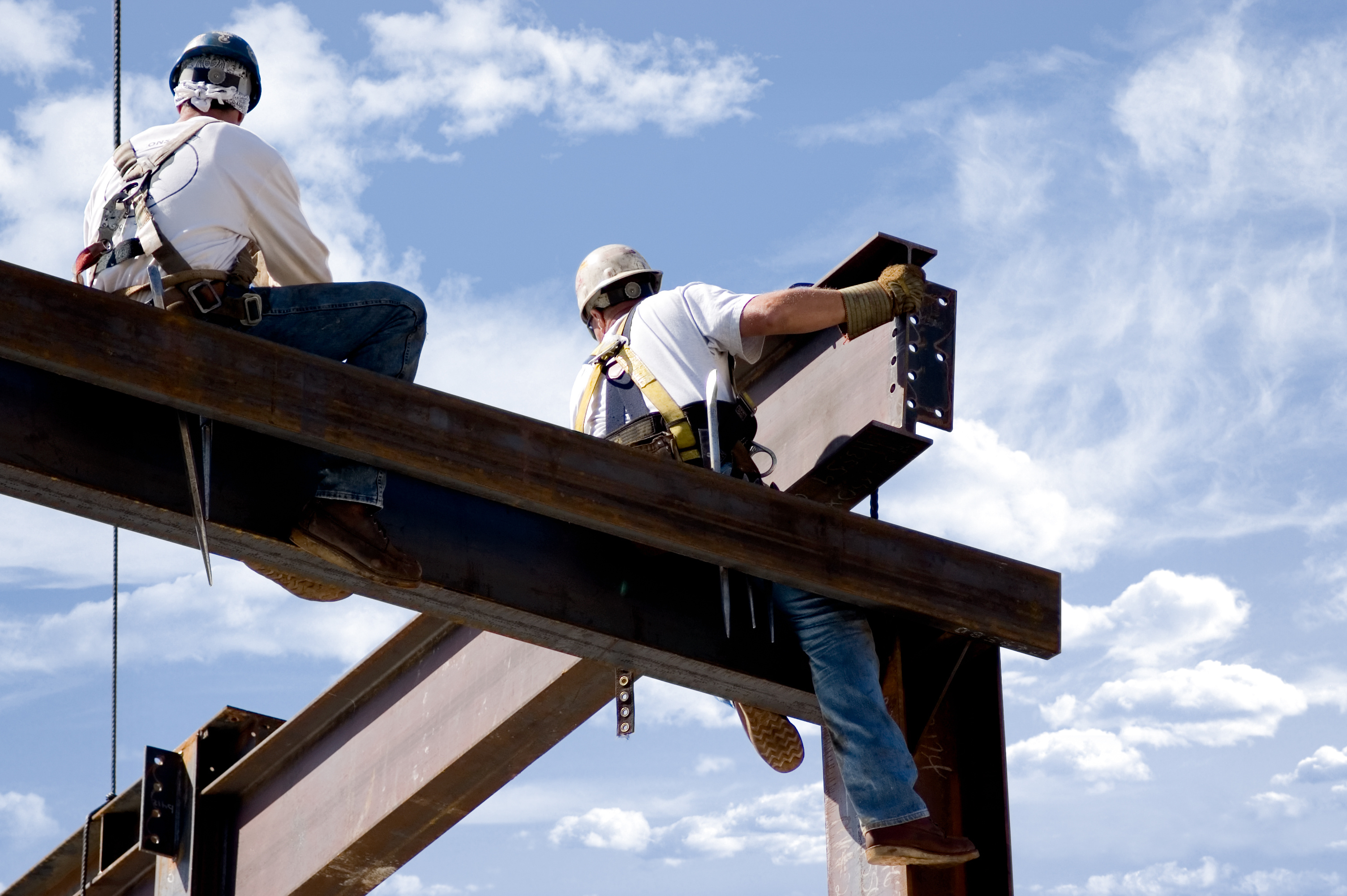 Two ironworkers atop the skeleton of a modern building. One man is positioning a very large beam while the other watches.